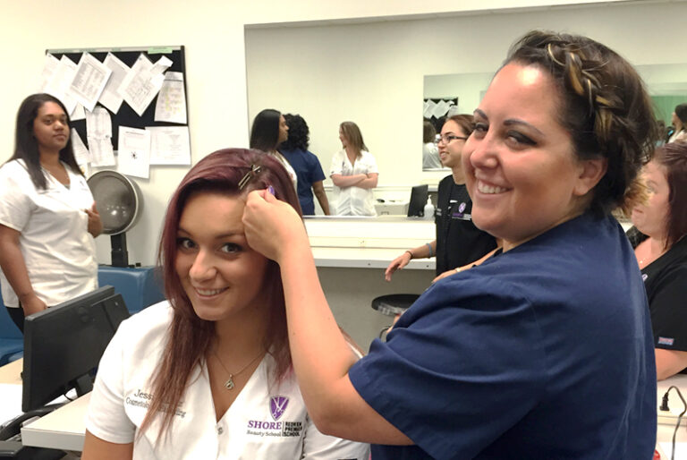 Beauty school students practicing hairstyling in a classroom setting with mirrors and smiles.