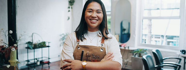 Smiling hairdresser in salon with apron and tools, standing confidently in stylish, well-lit space.