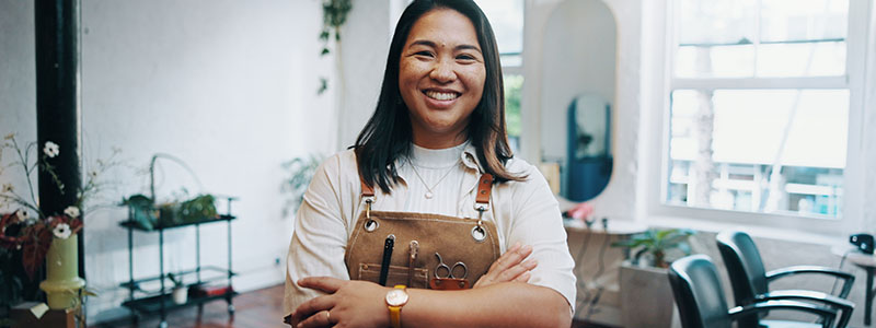 Smiling hairdresser in salon with apron and tools, standing confidently in stylish, well-lit space.
